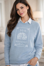 Brunette woman wearing a light blue 30A Mahjong Studio sweatshirt layered over a white collared shirt with pearl earrings in a soft neutral bedroom setting. Artwork © 2026 30A Mahjong Holdings LLC. All rights reserved.
