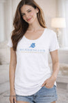 Brunette woman wearing a white 30A Mahjong Club T-shirt with I Survived My First Mahjong Lesson text in a soft neutral bedroom setting with pearl earrings and light makeup. Artwork © 2026 30A Mahjong Holdings LLC. All rights reserved.